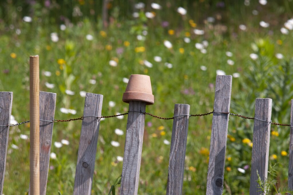 Rustikaler Holzzaun. Auf einer Latte steckt ein umgedrehter Blumentopf aus Ton. Im Hintergrund ist eine bunte Blumenwiese.
